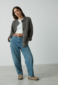Cardigan gris con relieve y grandes botones, llevado sobre una camiseta corta blanca y vaqueros de cintura alta azul. Acentuado con un cinturón rojo y zapatos beige.