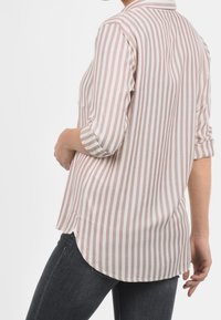 Persona che indossa una camicia a maniche lunghe, a righe verticali rosa e bianche con le maniche arrotolate e jeans scuri, girata leggermente di lato.