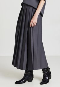 Donna che indossa una gonna midi plissettata scura con stivaletti in pelle nera sopra la caviglia caratterizzati da accenti di catena argentata, in piedi su un pavimento chiaro.