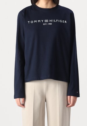 Person iført navyblå langærmet skjorte med teksten "Tommy Hilfiger Est. 1985" og logo, kombineret med lyse beige bukser mod ensfarvet baggrund.