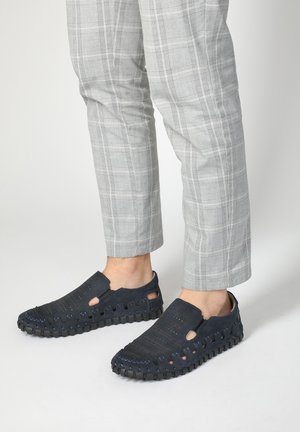 Chaussures noires à enfiler avec des trous découpés, semelle en caoutchouc texturée et accents en design tissé. Portées avec un pantalon à carreaux gris clair.