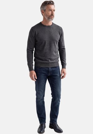 Man met grijs haar en baard, gekleed in een donkergrijze trui met ronde hals, blauwe spijkerbroek en zwarte veterlaarzen, staand en licht naar rechts gericht.