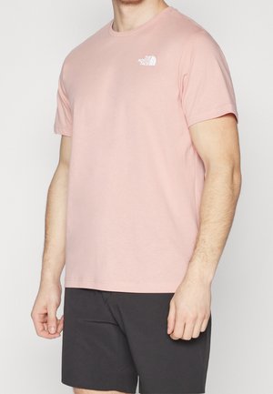 Mand iført lyserød The North Face kortærmet t-shirt og sorte shorts, vist fra hals til midt på låret mod ensfarvet baggrund.