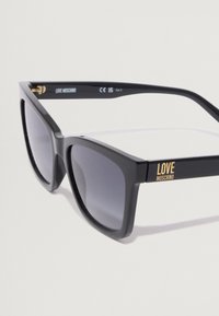 Ochelari de soare din plastic negru cu o formă pătrată îndrăzneață, lentile gri închis cu gradient și un logo "LOVE MOSCHINO" aurit pe brățări.