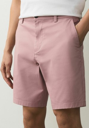 Man wearing light pink tailored shorts and a white shirt, with one hand in the pocket and the other relaxed by the side.