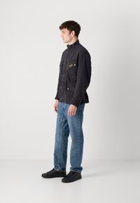 Casaco preto com cinto e dois bolsos no peito, pressionado com botões de metal, combinado com jeans de perna reta azul claro e ténis pretos.