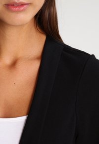 Černý blazer s hladkou texturou, se šálovým límcem a slim fit střihem. Pod blazerem je částečně vidět bílý top.