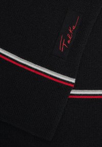 Tessuto a maglia nera con strisce rosse, bianche e grigie. Include un'etichetta rettangolare con "Falke" ricamato in rosso. Superficie testurizzata.