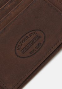 Brun ekte lær lommebok med debossert logo med "HILFIGER-NYC" og "est. 1985." Synlige kortspor og glatt tekstur.