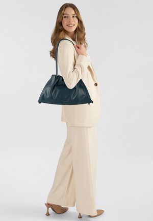 Borsa a tracolla in pelle color verde petrolio con un design morbido e arricciato e una singola tracolla, portata da una modella in un completo crema e tacchi beige.