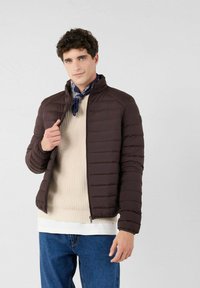 Bruine gewatteerde jas met hoge kraag, ritssluiting en elastische manchetten, gedragen over een beige trui en gelaagde blouse, met een blauwe sjaal.