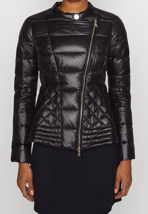 Femme portant une veste matelassée noire avec une fermeture éclair dorée asymétrique et un col à pression, associée à une jupe noire.