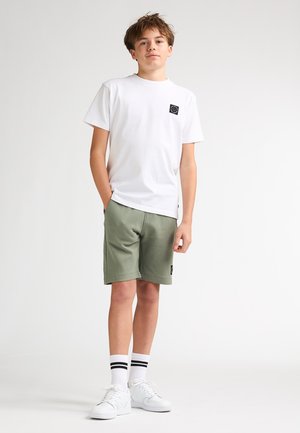 Tricou alb cu mâneci scurte, cu o etichetă logo neagră, asortat cu pantaloni scurți verde măsline. Colțurile pantalonilor scurți sunt ușor rotunjite. Adidași albi și șosete cu dungi.