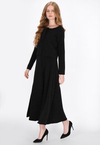 Rochie neagră midi cu mâneci lungi, din țesătură texturată cu sclipiri subtile. Corset ajustat și fustă fluidă, asortată cu pantofi cu toc înalt negri.