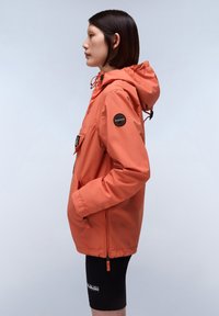Oranje lichtgewicht jack met een capuchon, met een voorzak, trekkoordsluiting en een zwart logo-embleem op de mouw. Zwarte shorts eronder.