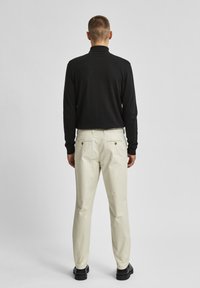 Černá košile s dlouhým rukávem spárovaná se světle béžovými kalhotami, které mají dvě zadní kapsy s kulatými kovovými knoflíky a texturovaným povrchem.