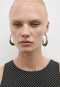 Modelka s světlými blond vlasy, oblečená v černém oděvu s bílými puntíky. Velké kovové kruhové náušnice podtrhují vzhled a doplňují minimalistický styl.