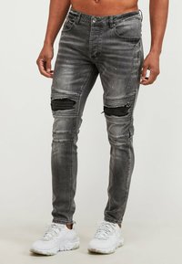 Vaqueros grises desgastados con detalles de rodillas rotas y tela texturizada. Combinados con zapatos deportivos blancos. Con múltiples bolsillos.