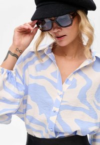 Vrouw met een zwarte hoed, grote tortoise-zonnebril, een blouse met een blauw-wit abstract patroon en die een infinity-tatoeage op haar linkerpols laat zien.