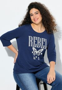 Marineblaue Langarm-T-Shirts mit einem "REBEL"-Grafik- und Schmetterlingsdesign; kombiniert mit Jeans aus Denim, die glitzernde Akzentdetails aufweisen.