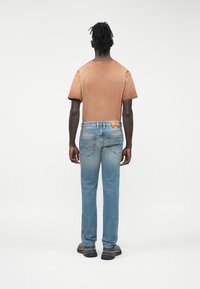 Camiseta marrón de manga corta, vaqueros de pierna recta azul claro y zapatillas negras, vistos desde atrás sobre un fondo blanco liso.