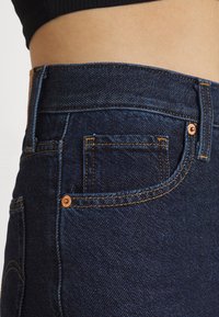 Jeans de mezclilla azul oscuro con una cintura alta, costuras naranjas visibles y un pequeño bolsillo trasero con un detalle de botón de cobre.