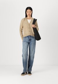 Beige strikket cardigan med V-hals og frontknapper, kombinert med blå rette jeans og svarte sko. Modellen bærer en svart veske.