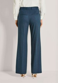 Vrouw in een hoog-taille, wijde blauwe broek en witte hoge hakken, staand met handen ontspannen langs de zijden.