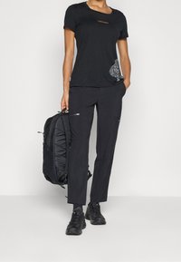 Czarna koszula z krótkim rękawem z subtelnym logo i wzorzystym dodatkiem, zestawiona z czarnymi spodniami z kieszeniami na zamek oraz czarnym plecakiem.