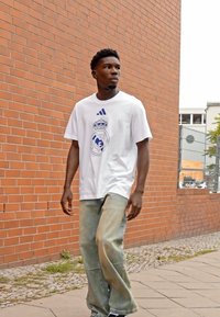 Hvid t-shirt med blåt Real Madrid-logo, parret med lyseblå løstsiddende jeans. Mand går på en sti nær en murstensvæg.