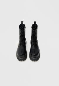 Botas de cano curto em pele preta com bico arredondado, sola grossa, fecho de atacadores à frente e fechos de correr laterais. Apresentam detalhes de costura na borda da sola.