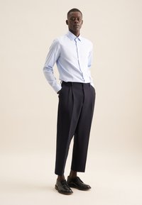 Camicia azzurro chiaro con bottoni, pantaloni blu navy a pieghe e scarpe nere stringate. Tessuto liscio, vestibilità su misura e elementi di design classici.