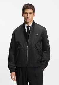 Black bomber jacket in smooth fabric with a front zipper, ribbed cuffs, and a distinctive logo patch. Features angled front pockets.