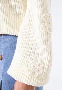 Cremfarbener, gestrickter Pulloverbund mit zwei gehäkelten Blumenpatches, getragen mit einem blauen Cordrock mit braunen Knöpfen.