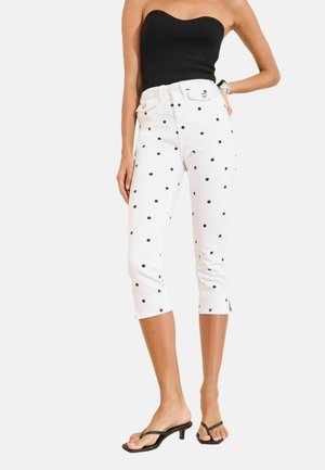 Vrouw draagt witte cropped broek met zwarte stippen, zwart strapless topje en zwarte sandalen met hakken, staand tegen een witte achtergrond.