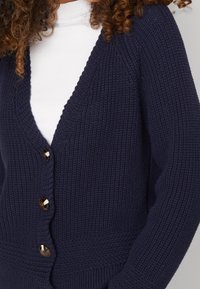 Cardigan lavorato a maglia blu navy con scollo a V, apertura frontale e bottoni dorati. Il tessuto testurizzato aggiunge profondità e calore al design.