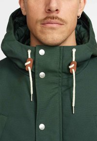 Groene waterdichte jas met een hoge kraag, zilveren drukknoopjes en verstelbare trekkoorden met leren accenten. Gestructureerde stof.