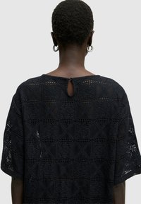 Schwarzes Strickoberteil mit geometrischen Mustern, kurzen Ärmeln und einem Rückenschlitz mit Knopfdetails. Gittertextur im gesamten Design.