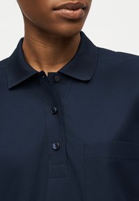 Κοντινό πλάνο ενός ατόμου που φοράει σκούρο μπλε πόλο με τρία κουμπιά και τσέπη στο στήθος με διακριτικό λογότυπο κροκόδειλου.