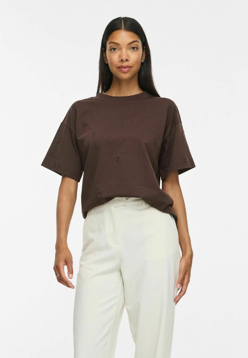Femme aux longs cheveux foncés porte un haut marron ample à manches courtes et un pantalon blanc taille haute, debout devant un fond blanc uni.