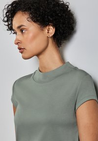 Krátký rukáv top v měkké, tlumené zelené tkanině. Má vysoký, zaoblený výstřih a hladkou strukturu. Viditelné jsou zesílené švy na ramenech.
