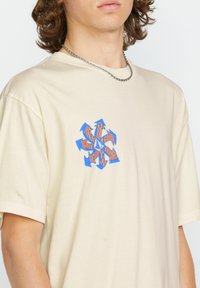 Camiseta de algodón color crema con un gráfico geométrico en azul y naranja en el pecho. Mangas cortas y un diseño de corte relajado.