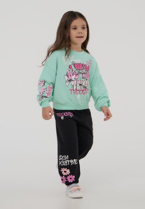 Felpa oversized verde menta con grafiche rosa e pantaloni della tuta neri con testo bianco e rosa. Scarpe chiare con accenti pastello.