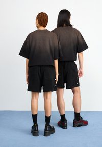 Zwarte gradient t-shirts met oversized pasvorm, bijpassende zwarte badstof korte broeken. Beide hebben zakken en een textuur materiaal. Zwarte schoenen met rode accenten.