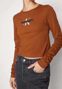 Hnědý dlouhý rukáv crop top s bílým logem "Calvin Klein Jeans" a černým akcentem. V kombinaci s tmavými džíny.
