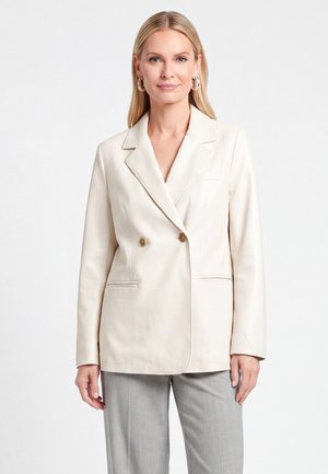Blonde vrouw die een crèmekleurig dubbelgeknoopt blazer draagt met gouden knopen en lichtgrijze broek, staand voor een eenvoudige witte achtergrond.