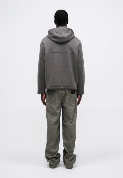 Hanorac gri cu o croială lejeră și glugă cu șnur, asortat cu pantaloni largi, de culoare verde estompată. Design minimalist, fără modele vizibile.