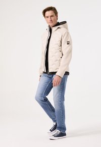 Gevoerde lichtgewicht jas in crème met een gestructureerd oppervlak, een zwarte capuchon en ritszakken. Gecombineerd met denim jeans en sneakers.