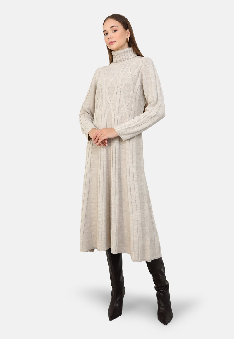 Robe tricotée beige avec un col haut, des manches longues, des motifs en cable et un ourlet évasé, associée à des bottes noires jusqu'aux genoux.