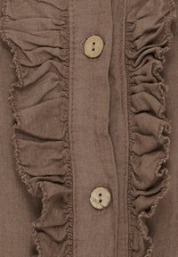 Brun linned skjorte med en central flæsepanel og fire runde træknapper. Teksturen er blød, med synlige syninger.